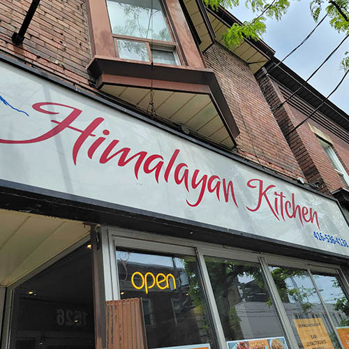 Himalayan Kitchen has a white wide sign with cursive style writing in red that says Himalayan Kitchen. Theyre open sign lights up in a neon yellow hue