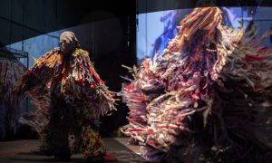an unrecognizable figure twirls around on stage wearing layers and layers of long tassels made of various colour fabric. Projections overlay the performer.