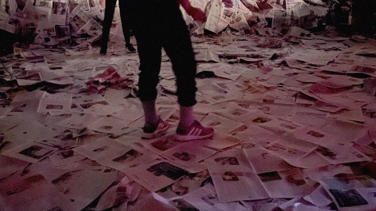 A blurry photo of a dancer in sneakers standing on a stage and wall covered in newspapers. This image is from “Okay, you can stop now”.