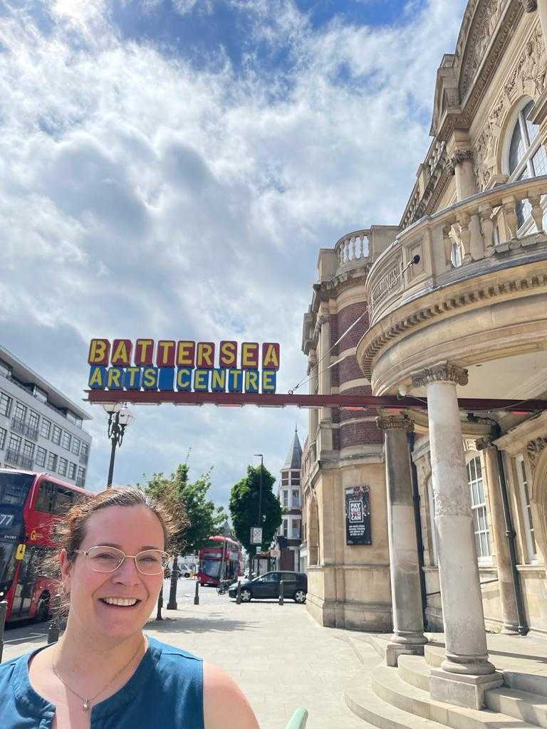 Kelsie smiles in front of Battersea which is a heritage building with a sign that sticks out a bit comically, that reads BATTERSEA Arts Centre. Kelsey wears glasses and smiles in a blue dress.