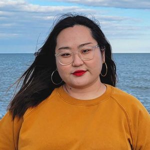 Emily is a Korean woman with long brown hair. She is smiling in front of waters, wearing a red lipstick, hoop earrings, and a mustard sweater.