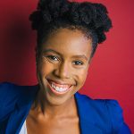 Playwright Kanika Ambrose smiles brightly in front of a red backdrop wall. She has her hair tied up and is wearing a bright blue blazer and a white shirt.