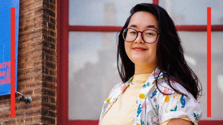 Nicole is a mixed-raced person of east asian decent. She has dark black hair that is blowing in the wind. She wears glasses, a yellow turtleneck and a fun confetti patterned shirt overtop.