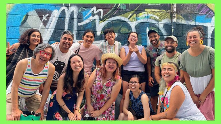 participants of the Digital Creators Lab gather together in front of an energetic blue graffiti wall for a photo. Everyone is in summery clothes, smiling.