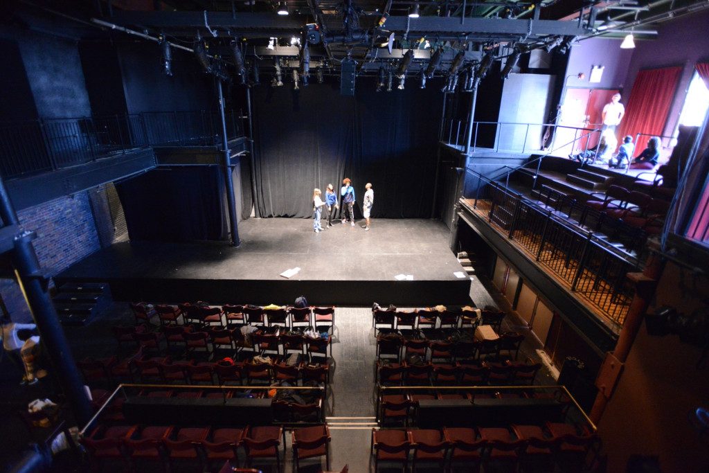 photo of the stage area taken from the second floor balcony, there are seats in the audience on either side of an aisle that are dark red and the stage is raised approx. 3 feet with a section to the left that has a second floor overhang, there is also a side seating area on the second floor to the right.