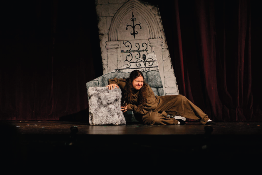 Lilia in front of a stone structure on the stage in a brown costume with a scrunched face.